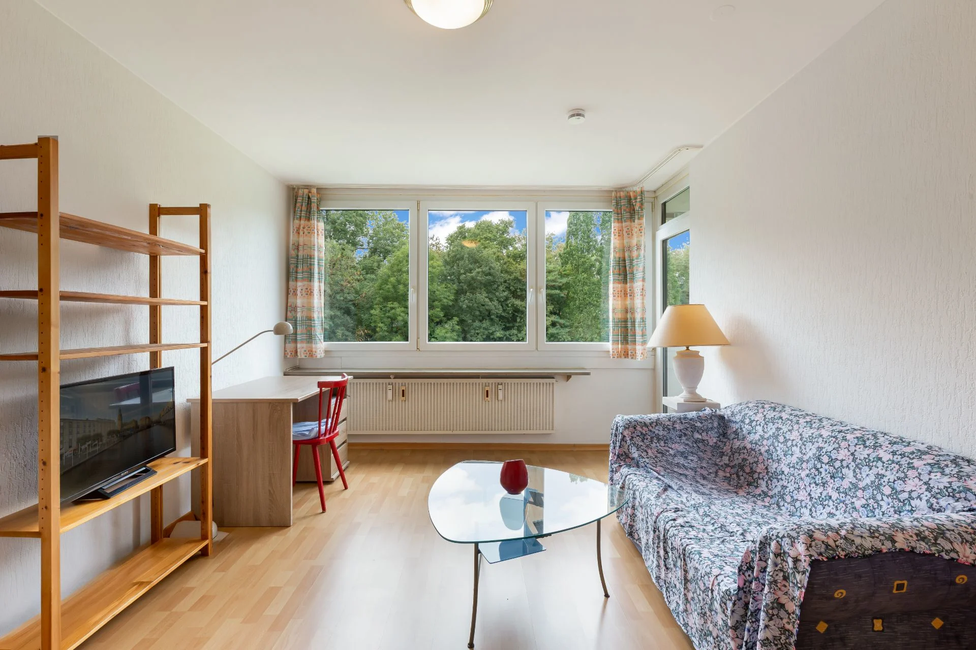 Wohnzimmer mit Sofa, Glastisch, Regal mit Fernseher, Schreibtisch und großem Fenster mit Blick ins Grüne.