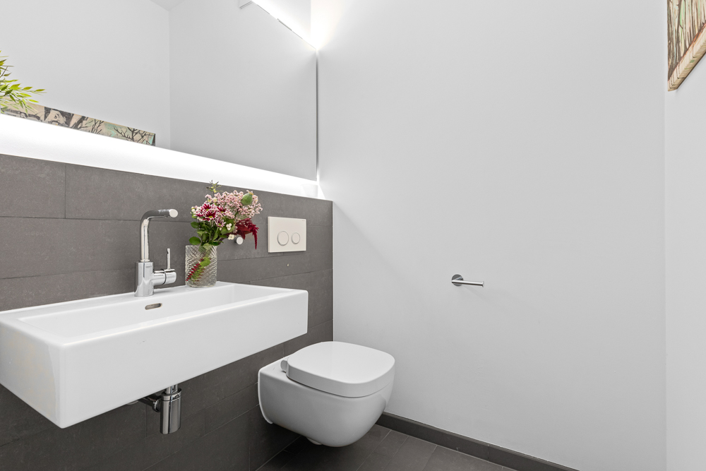 Modernes Badezimmer mit weißem Waschbecken, Wandtoilette und Vase mit Blumen.