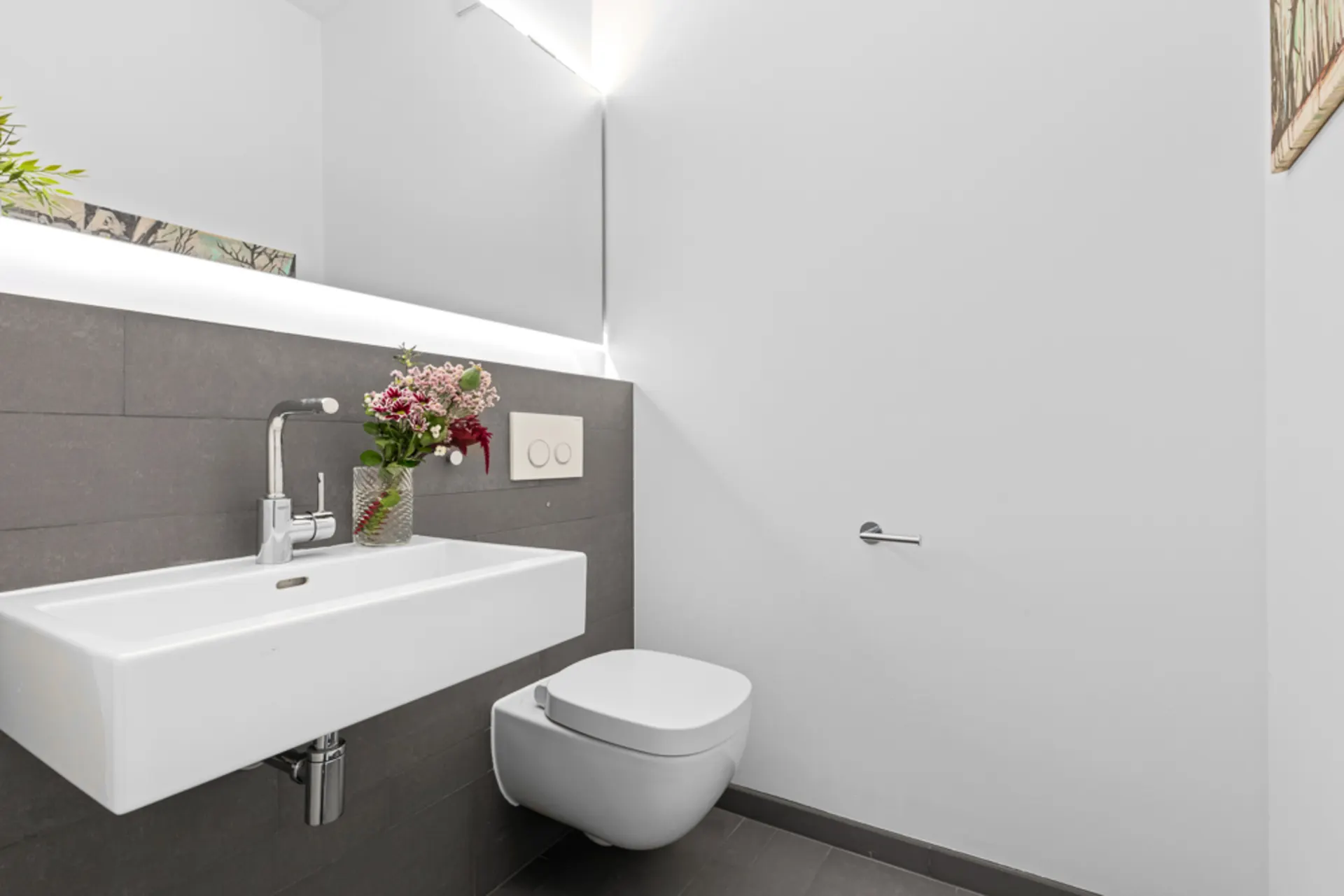 Modernes Badezimmer mit weißem Waschbecken, Wandtoilette und Vase mit Blumen.