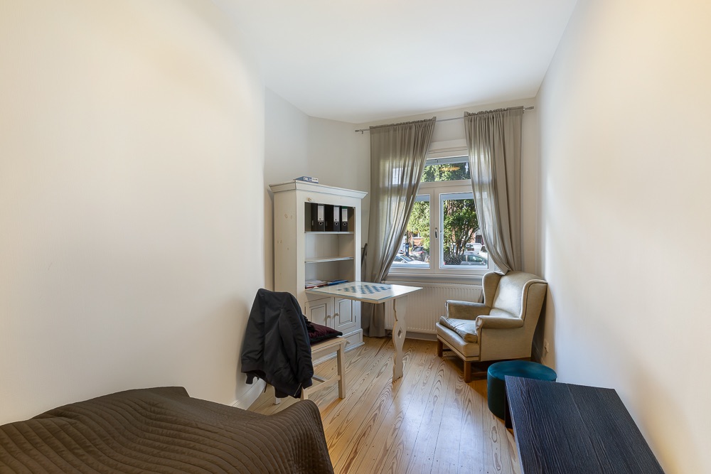 Helles Zimmer mit Sessel, Tisch, Regal, Bett, Fenster mit Vorhängen und Parkettboden.