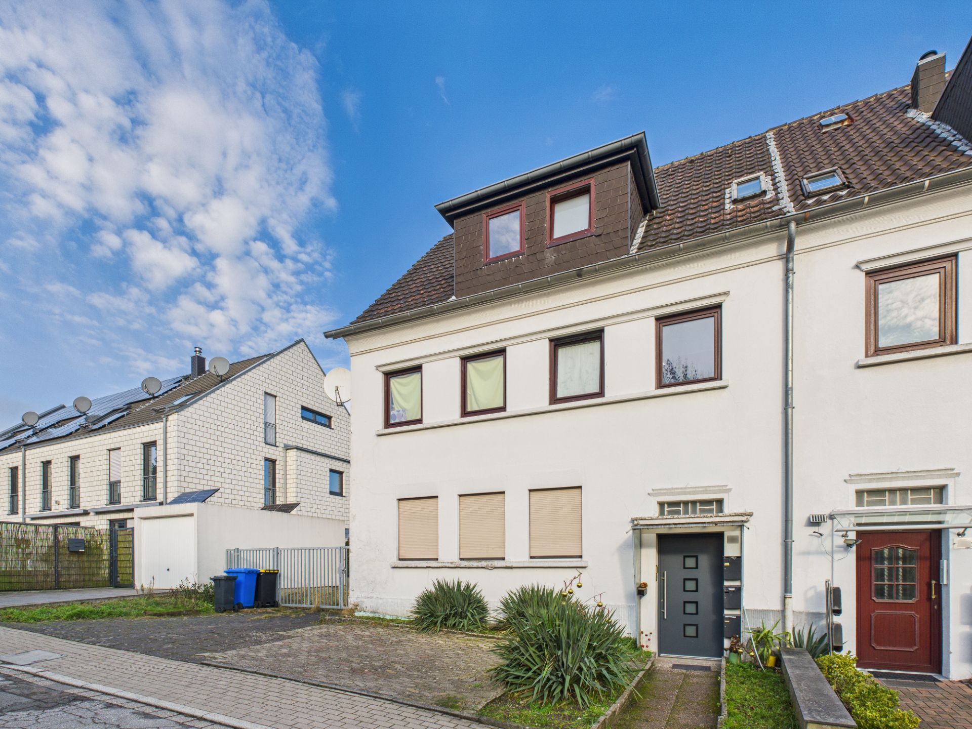 Weiße Reihenhausfassade mit mehreren Fenstern, schwarzer Tür und Vorgartenpflanzen bei Tag.