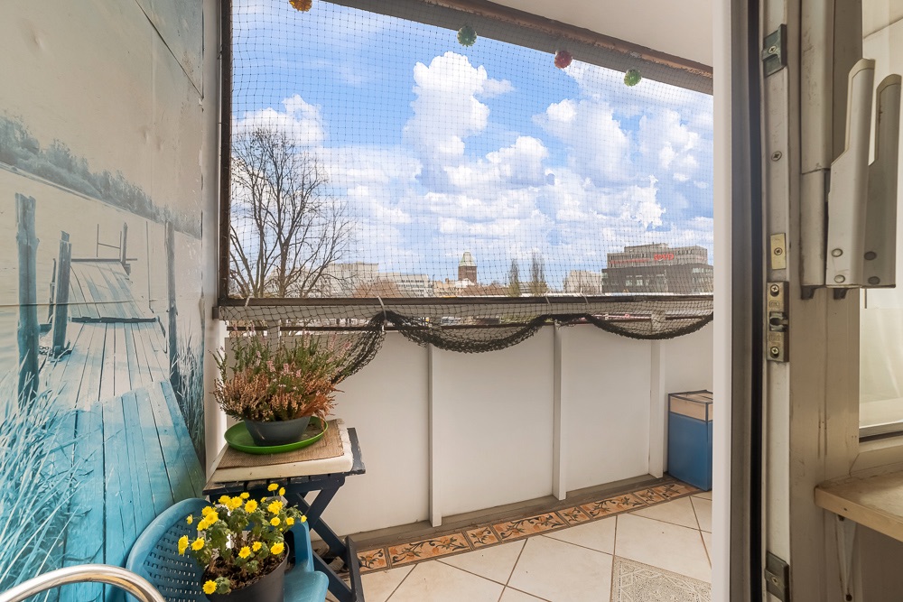 Kleiner Balkon mit Netz, Stuhl, Blumen, Tisch und Ausblick auf Gebäude bei blauem Himmel.