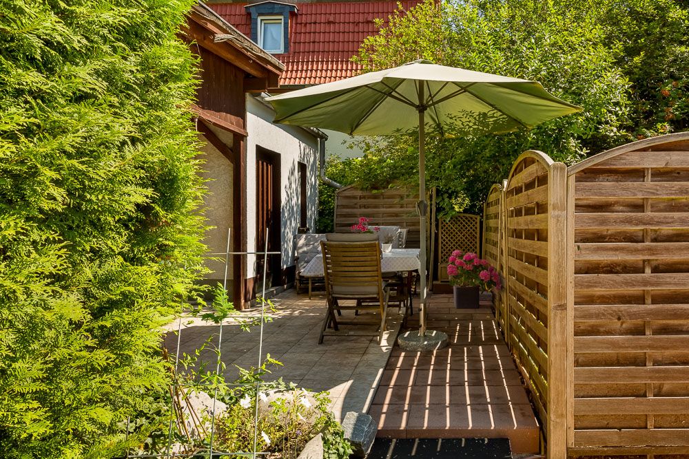 Terrasse mit Tisch, Stühlen, Sonnenschirm und Holzzaun, umgeben von Pflanzen.