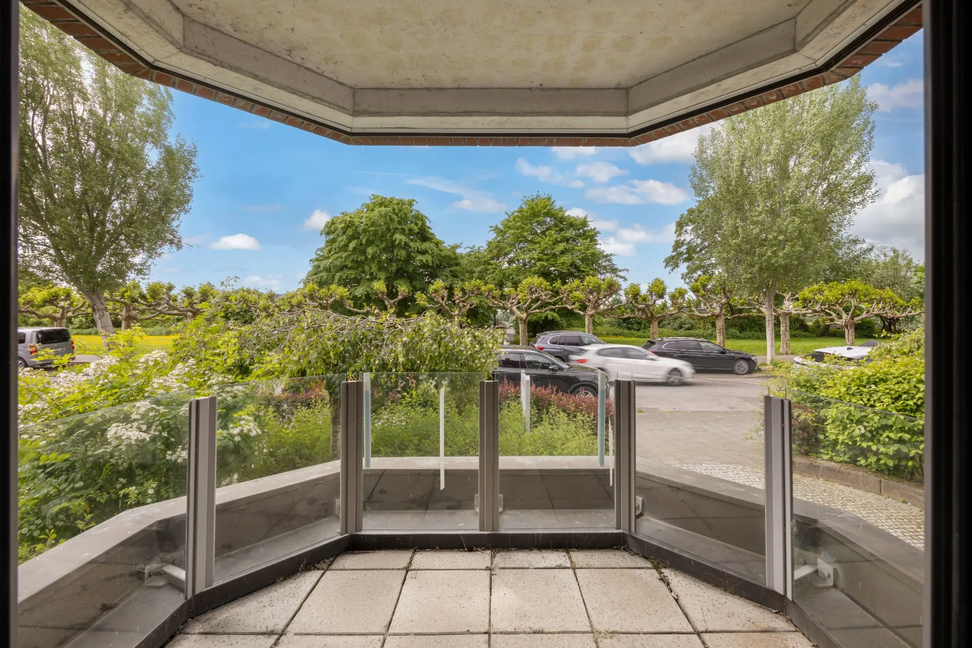 Kleiner Balkon mit Glasgeländer, Blick auf Straße, parkende Autos und grüne Bäume.