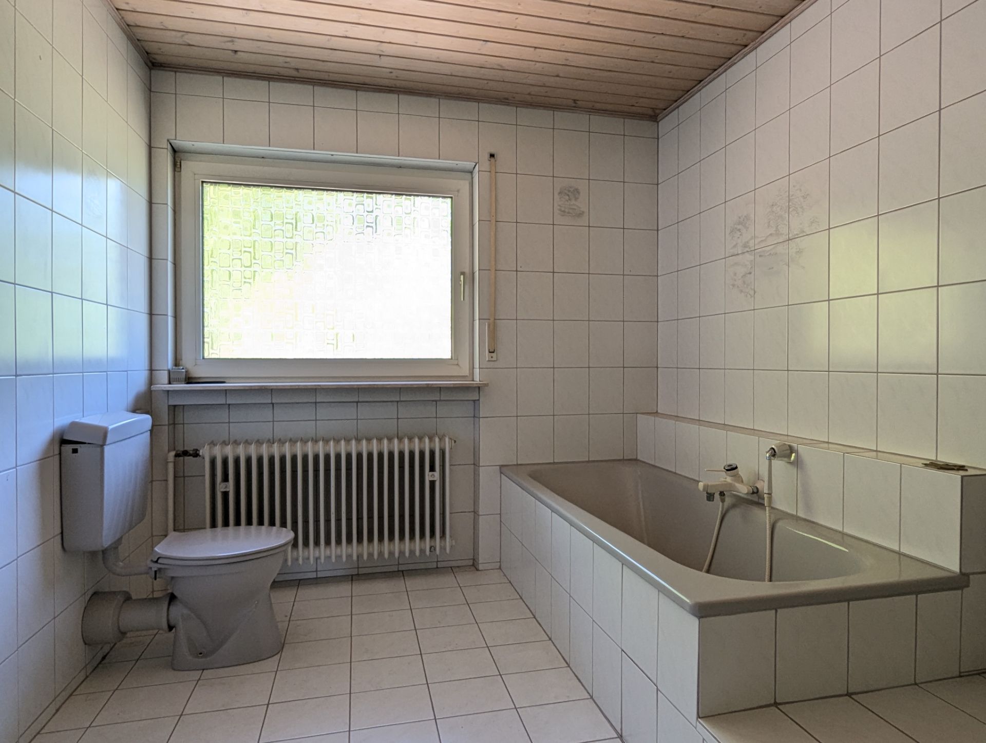 Helles Badezimmer mit Badewanne, Toilette, Fenster und Heizkörper, gefliest und Holzdecke.
