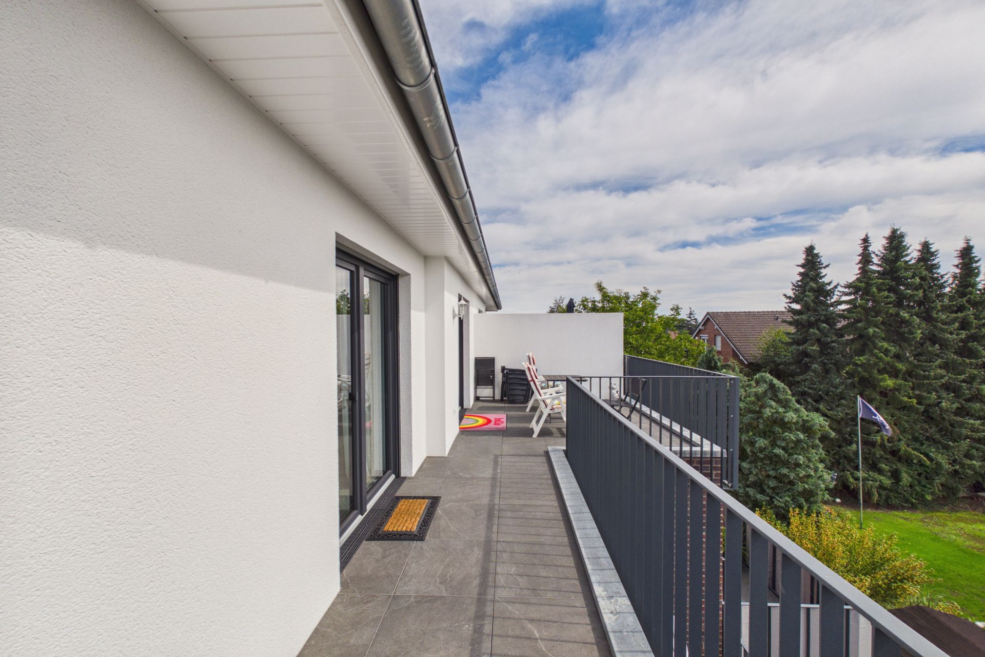 Großer Balkon mit grauem Boden, weißen Wänden und Gartenblick, Stühle im Hintergrund.