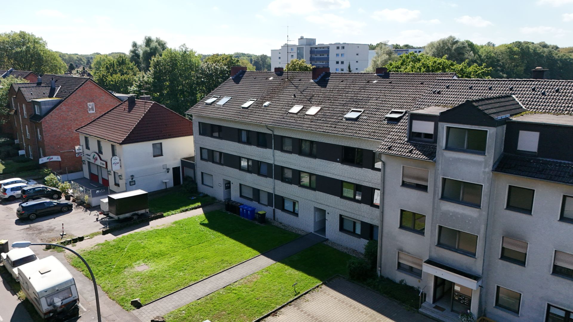 Mehrfamilienhaus mit drei Etagen, Vorgarten und parkenden Autos an der Straße.