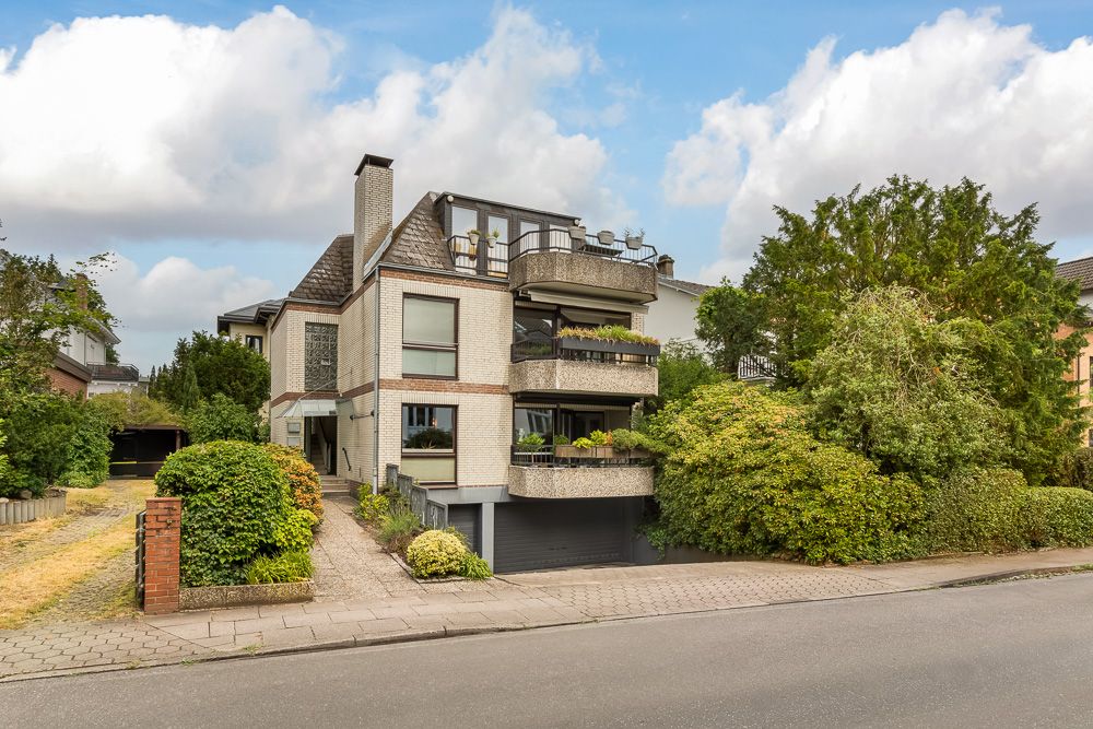 Modernes Wohnhaus mit mehreren Balkonen, Garage und viel Grün vor dem Haus.
