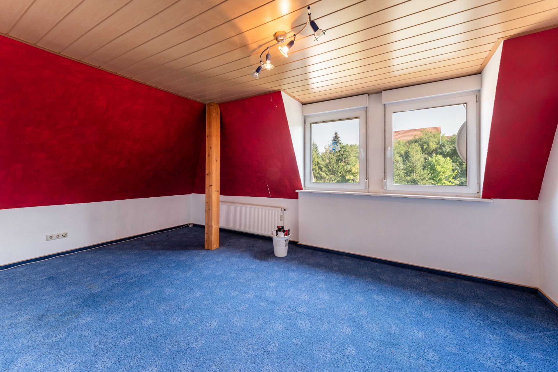 Leeres Zimmer mit blauem Teppich, roten Wänden, Holzdecke und zwei Fenstern.