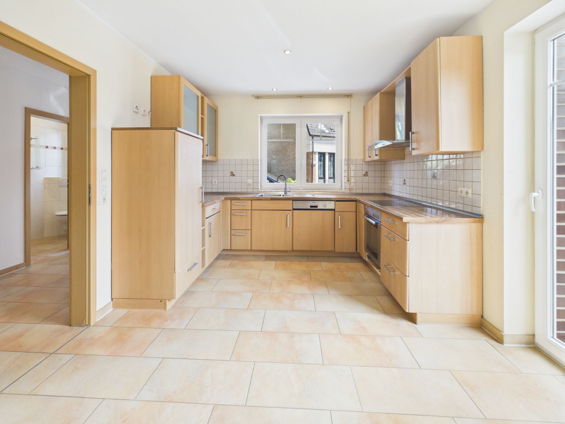 Helle Einbauküche mit Holzschränken, Fliesenboden und Fenster, angrenzend an ein weiteres Zimmer.