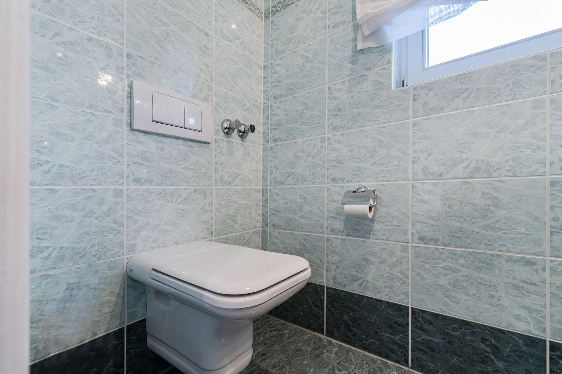 Moderne Toilette mit blauen Fliesen und Fenster, Toilettenpapier an der Wand.