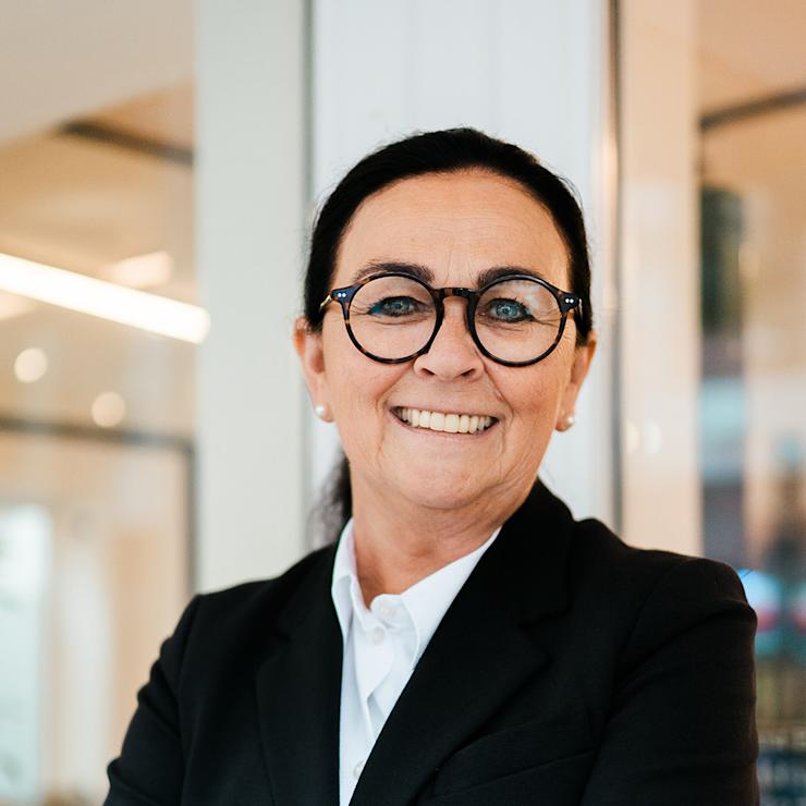 Frau mit Brille und schwarzem Blazer steht lächelnd mit verschränkten Armen in einem Raum.