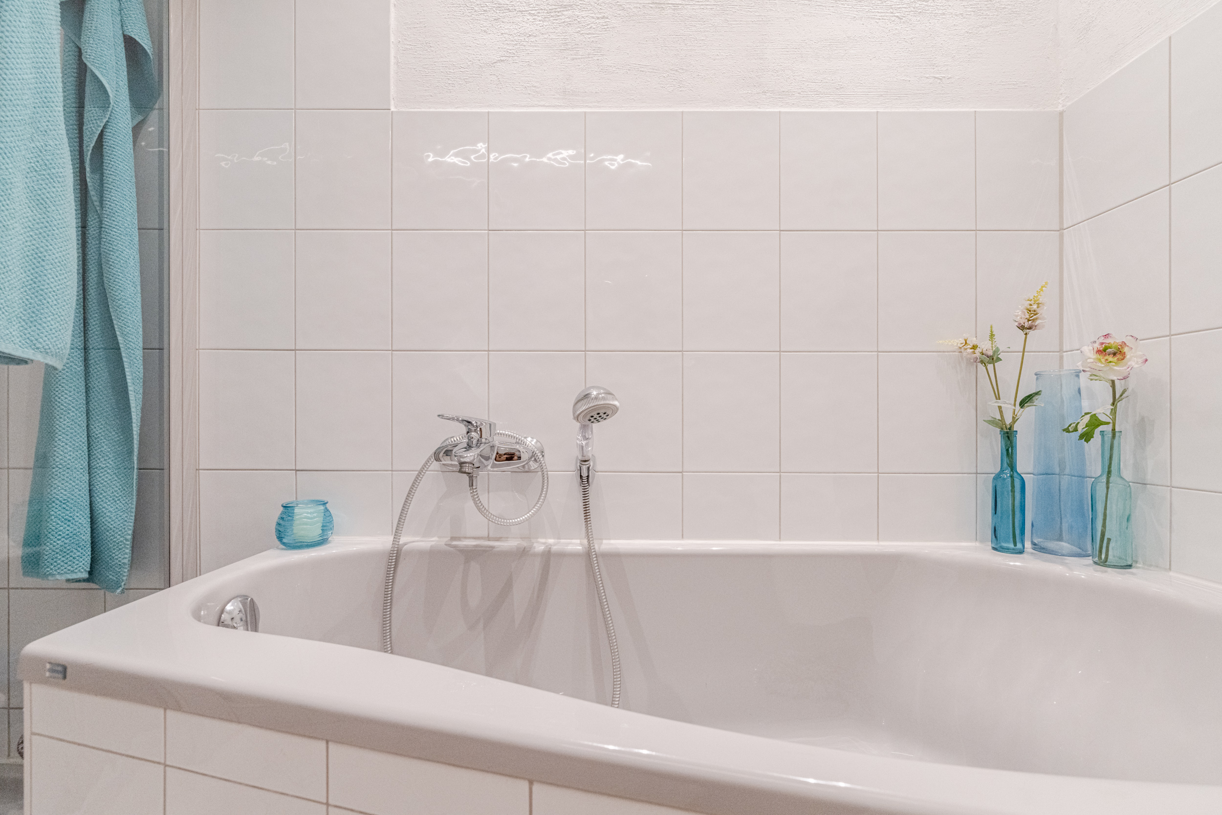 Weiße Badewanne mit Duscharmatur, blauen Handtüchern und Blumenvasen auf einer Fliesecke.