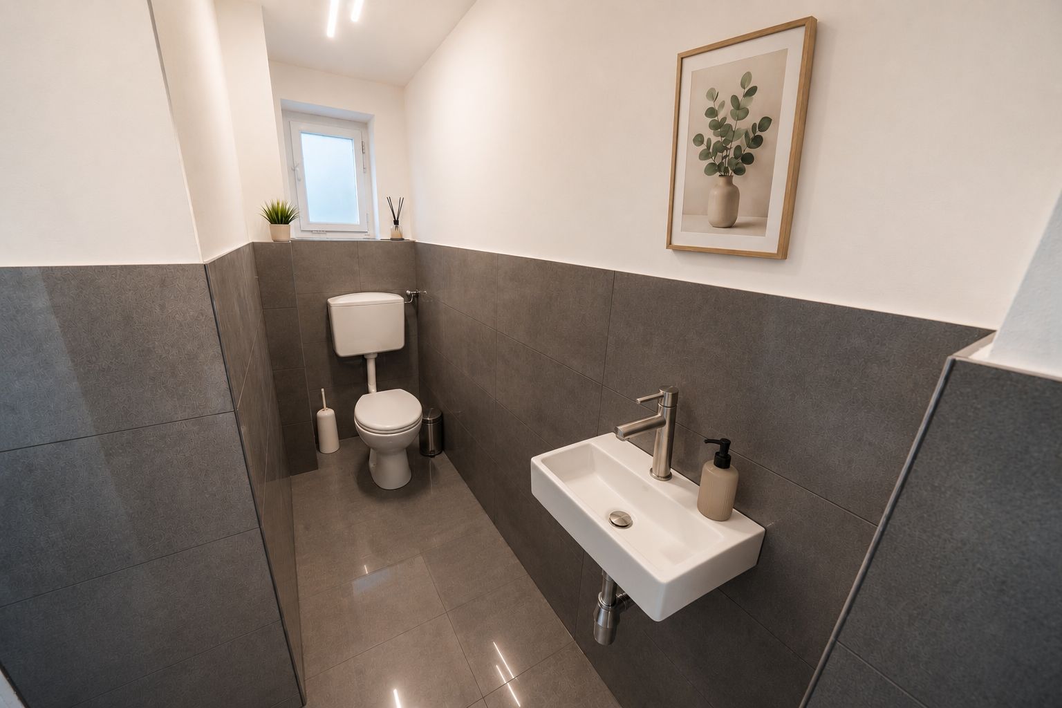 Modernes WC mit kleinem Waschbecken, grauen Fliesen und Pflanzenbild an der Wand.