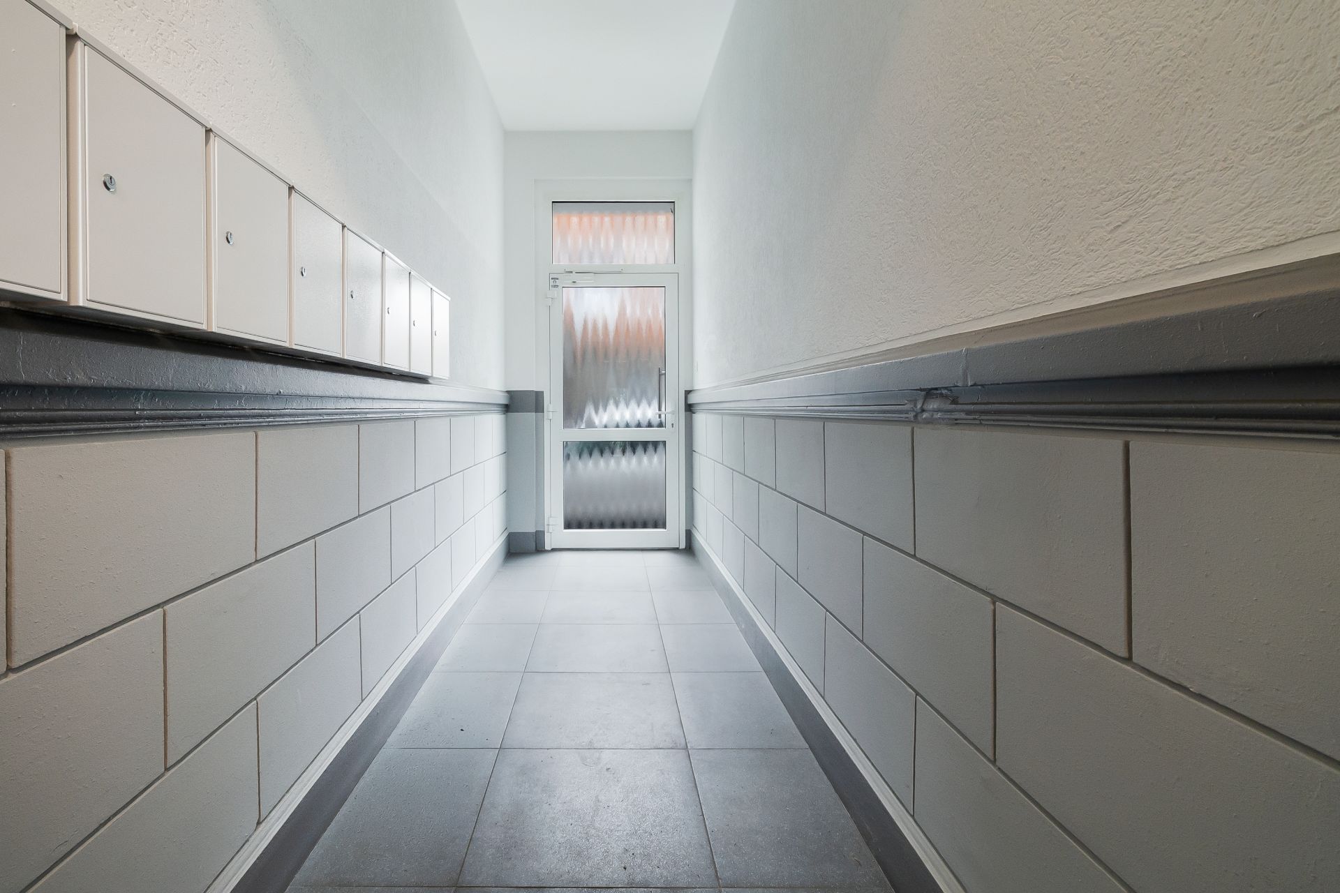 Heller Flur mit weißen Fliesen und Briefkästen an der Wand, am Ende eine Glastür.
