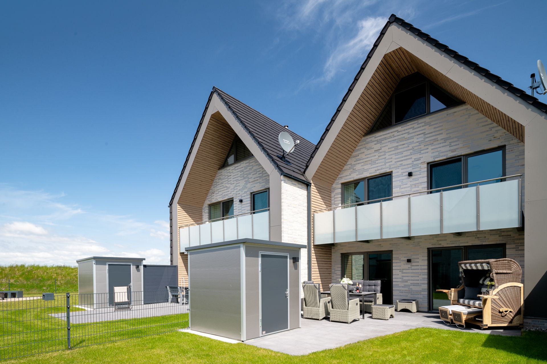 Zwei moderne Doppelhaushälften mit Balkon, Terrassenmöbeln und Garten auf einer Wiese.