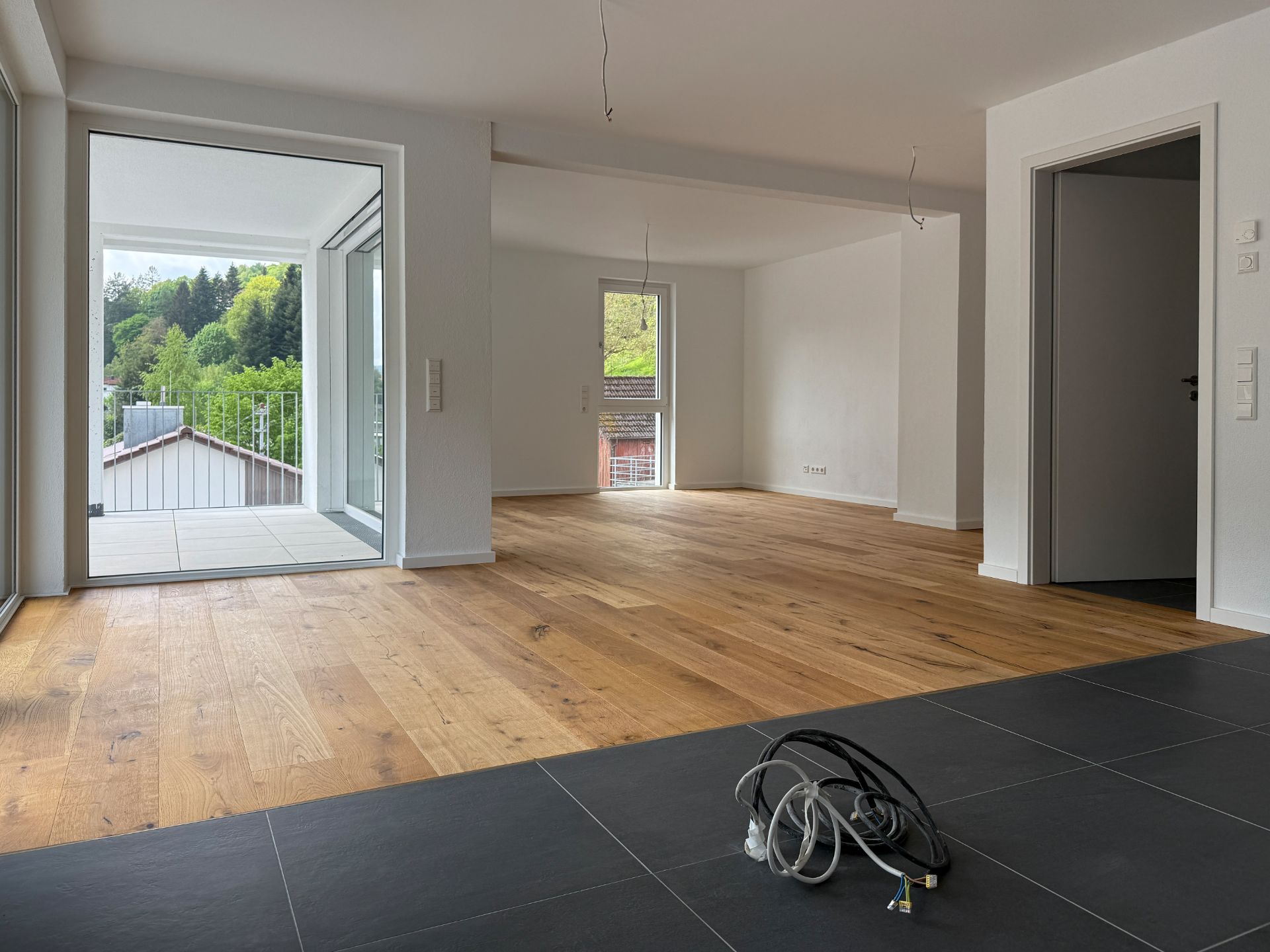Heller, leerer Wohnraum mit Holzboden, großer Fensterfront und Blick auf einen Balkon und grüne Bäume.
