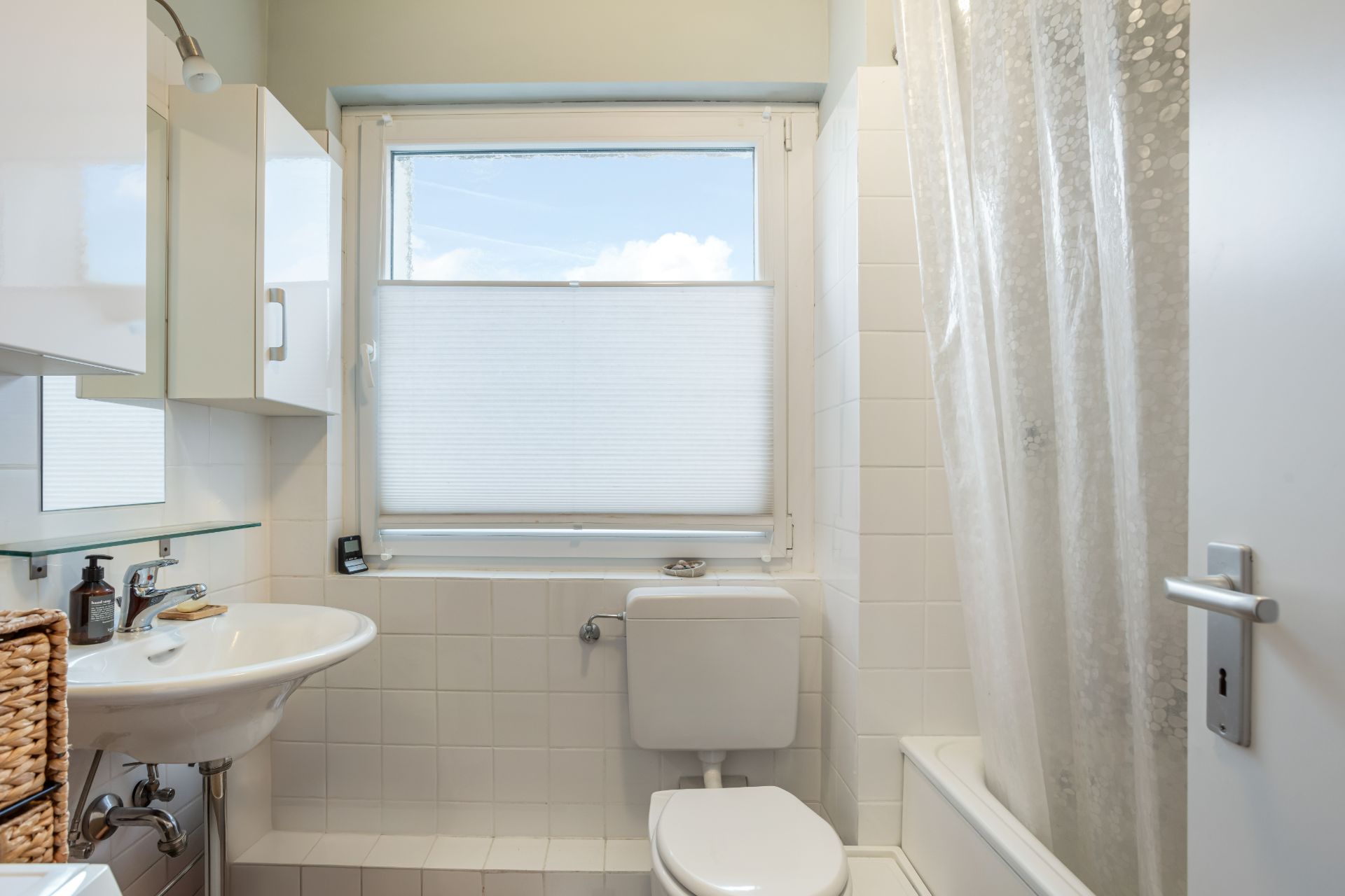 Kleines Badezimmer mit Waschbecken, Toilette, Fenster und Badewanne mit Duschvorhang.