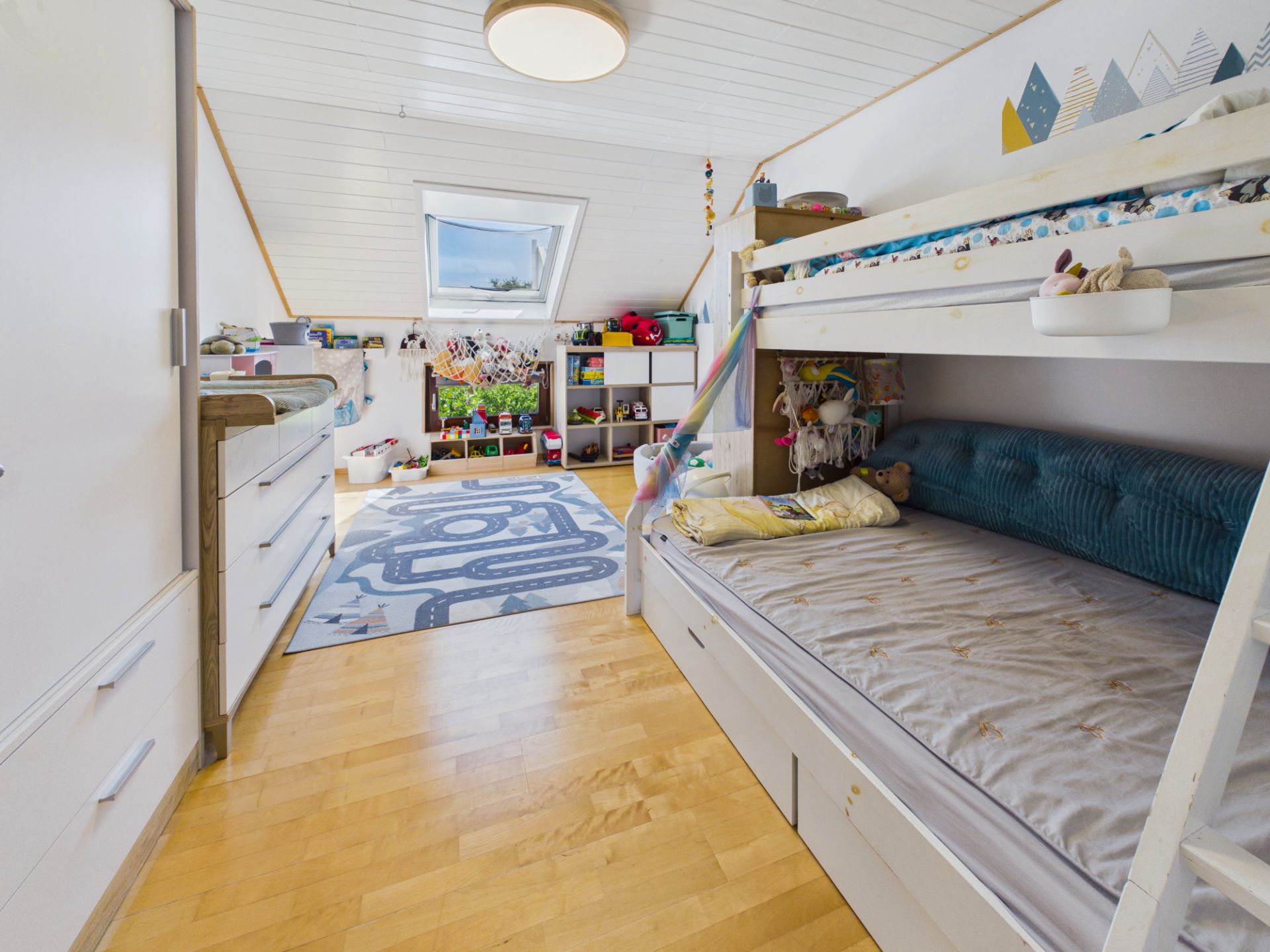 Helles Kinderzimmer mit Etagenbett, Holzfußboden, Spielteppich und Regalen unter einem Dachfenster.