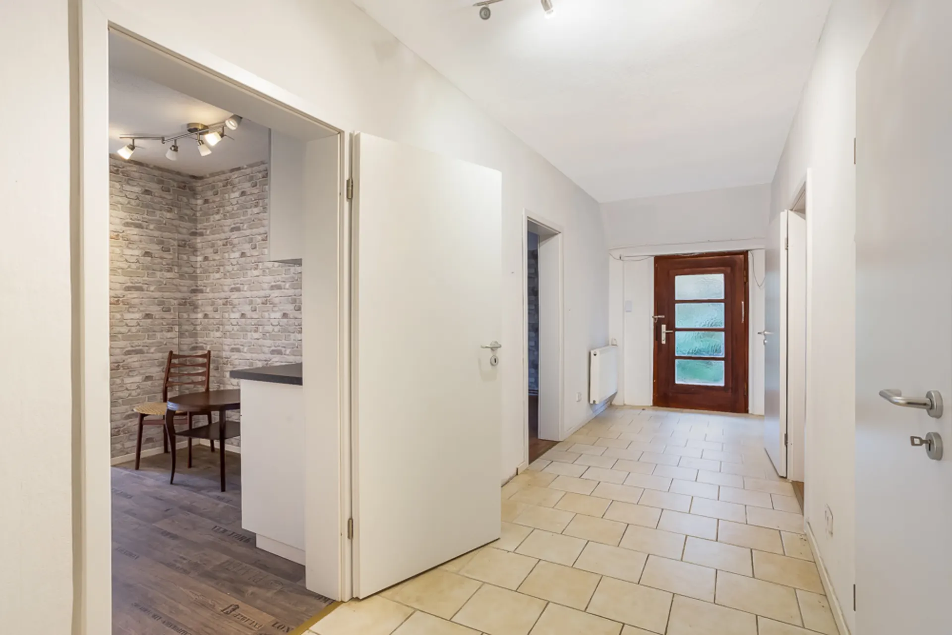 Heller Flur mit gefliestem Boden, mehreren Türen und Blick in ein Zimmer mit gemusterter Tapete.