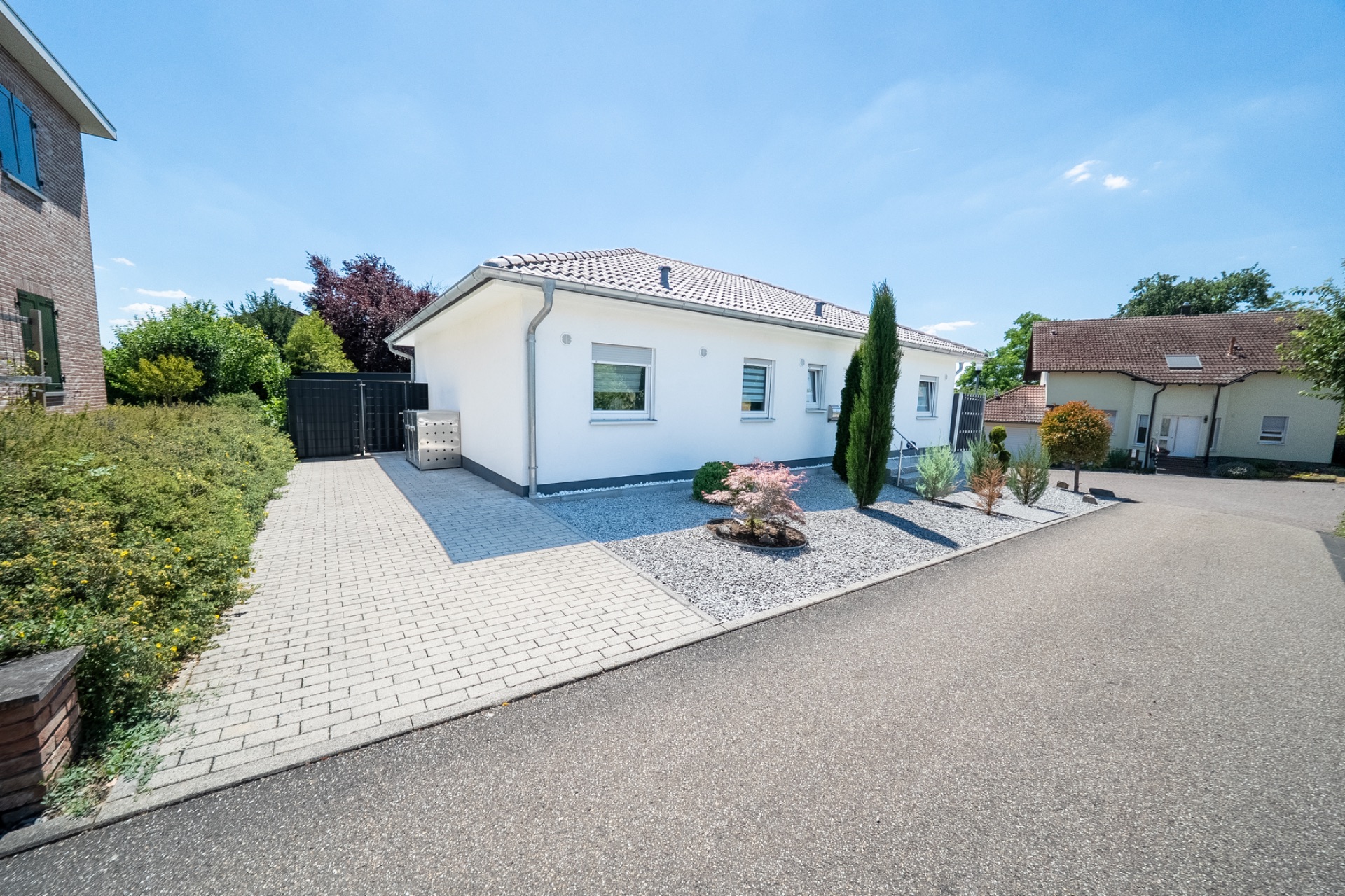 Modernes, weißes Wohnhaus mit Ziegeldach und Vorgarten, neben gepflasterter Einfahrt.