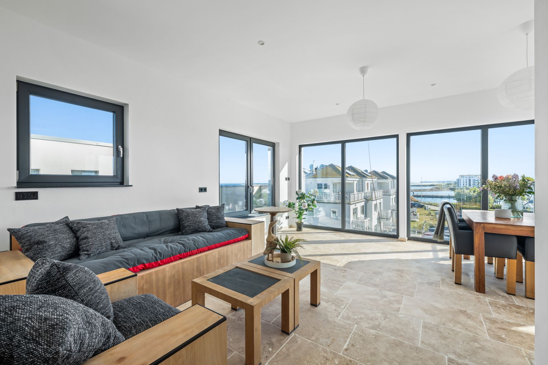 Helles Wohnzimmer mit Sofa, Tisch, Essbereich und großen Fenstern mit Blick nach draußen.