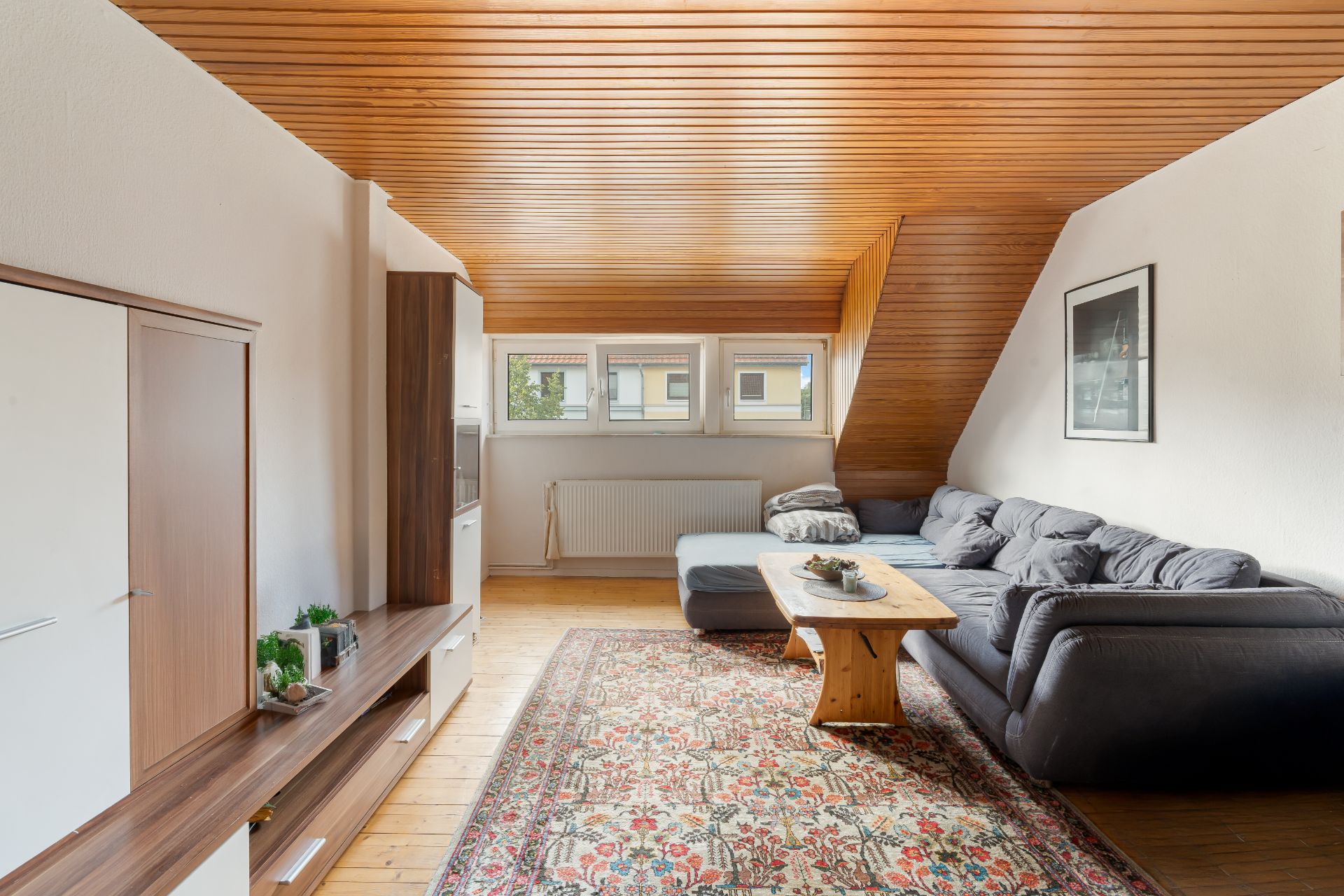 Wohnzimmer mit grauem Sofa, Holztisch, Teppich und Holzdecke, Fenster an der Stirnseite.