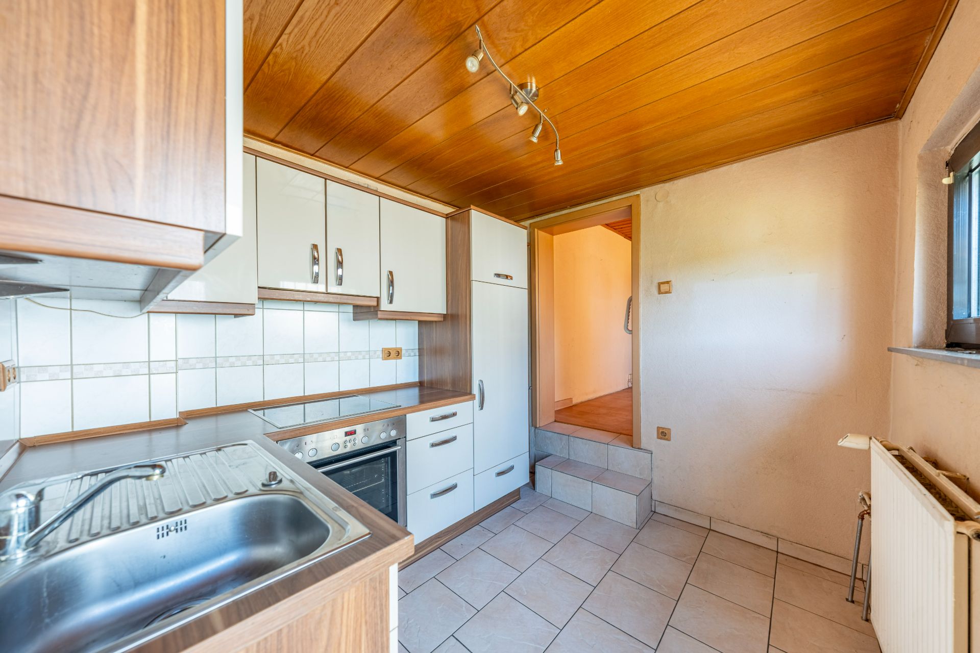 Helle Küche mit Holzdecke, weißen Schränken, Spüle, Herd und Fliesenboden.