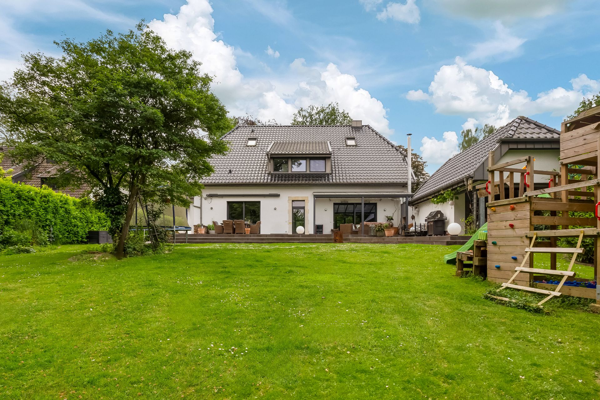 Rückansicht eines Hauses mit Terrasse, Garten, Baum und Holzspielplatz.