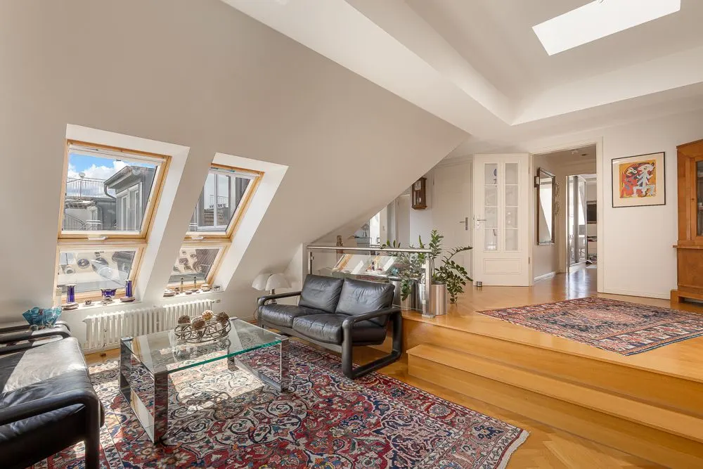 Helles Wohnzimmer mit Dachfenstern, zwei Ledersofas, Glastisch und Teppich auf Holzboden.