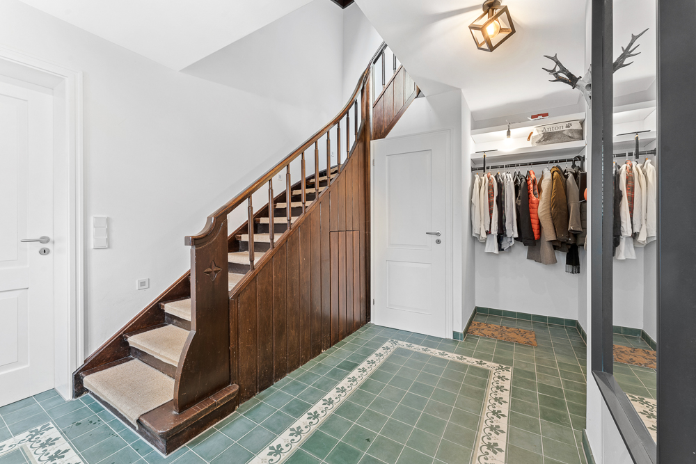 Flur mit grünen Fliesen, Holztreppe, Garderobe mit Kleidung und weißer Tür.