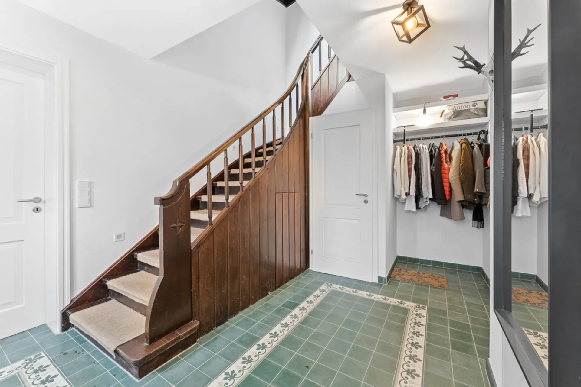 Flur mit grünen Fliesen, Holztreppe, Garderobe mit Kleidung und weißer Tür.