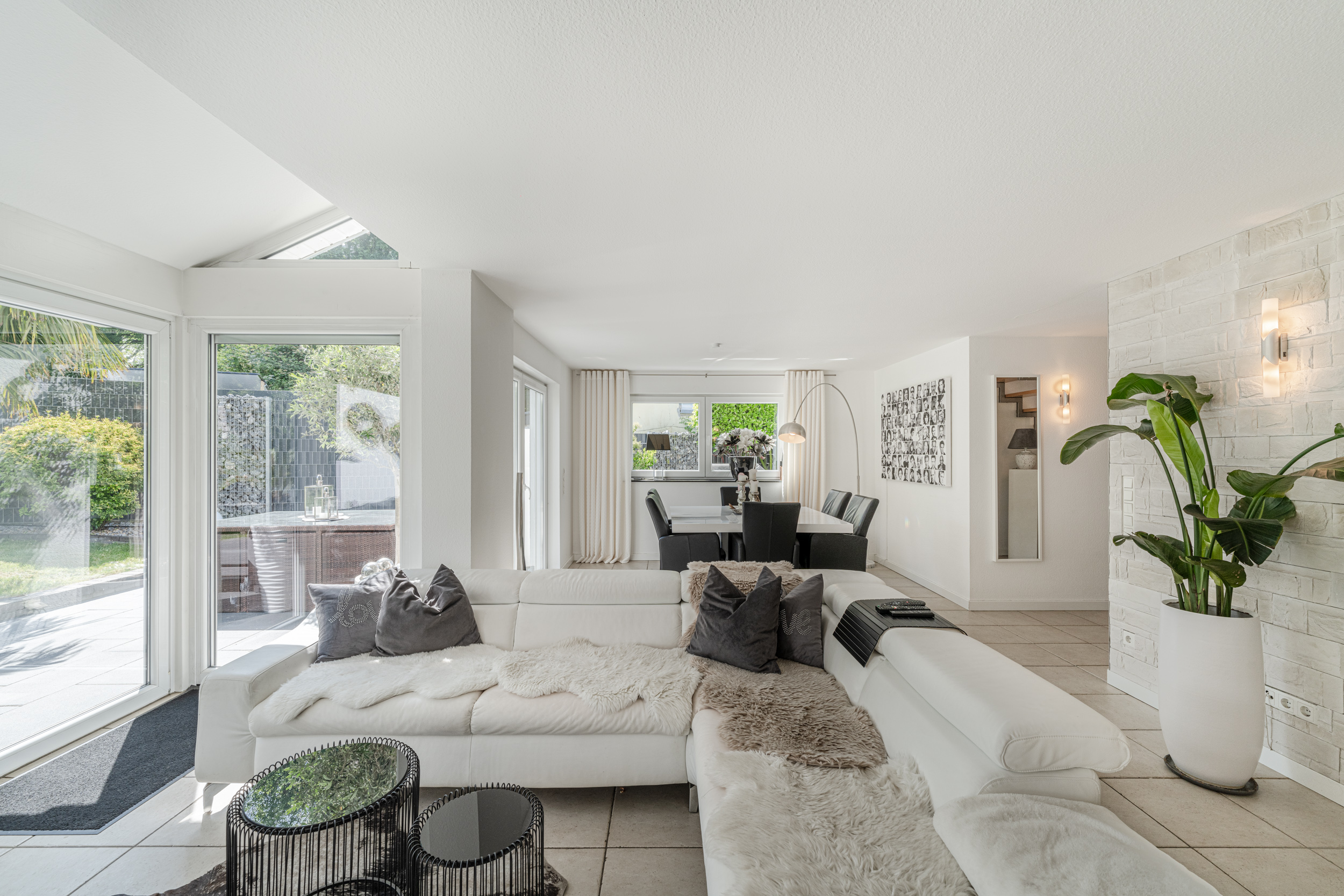 Helles Wohnzimmer mit weißem Sofa, Glastischen, Essbereich und großen Fenstern zum Garten.