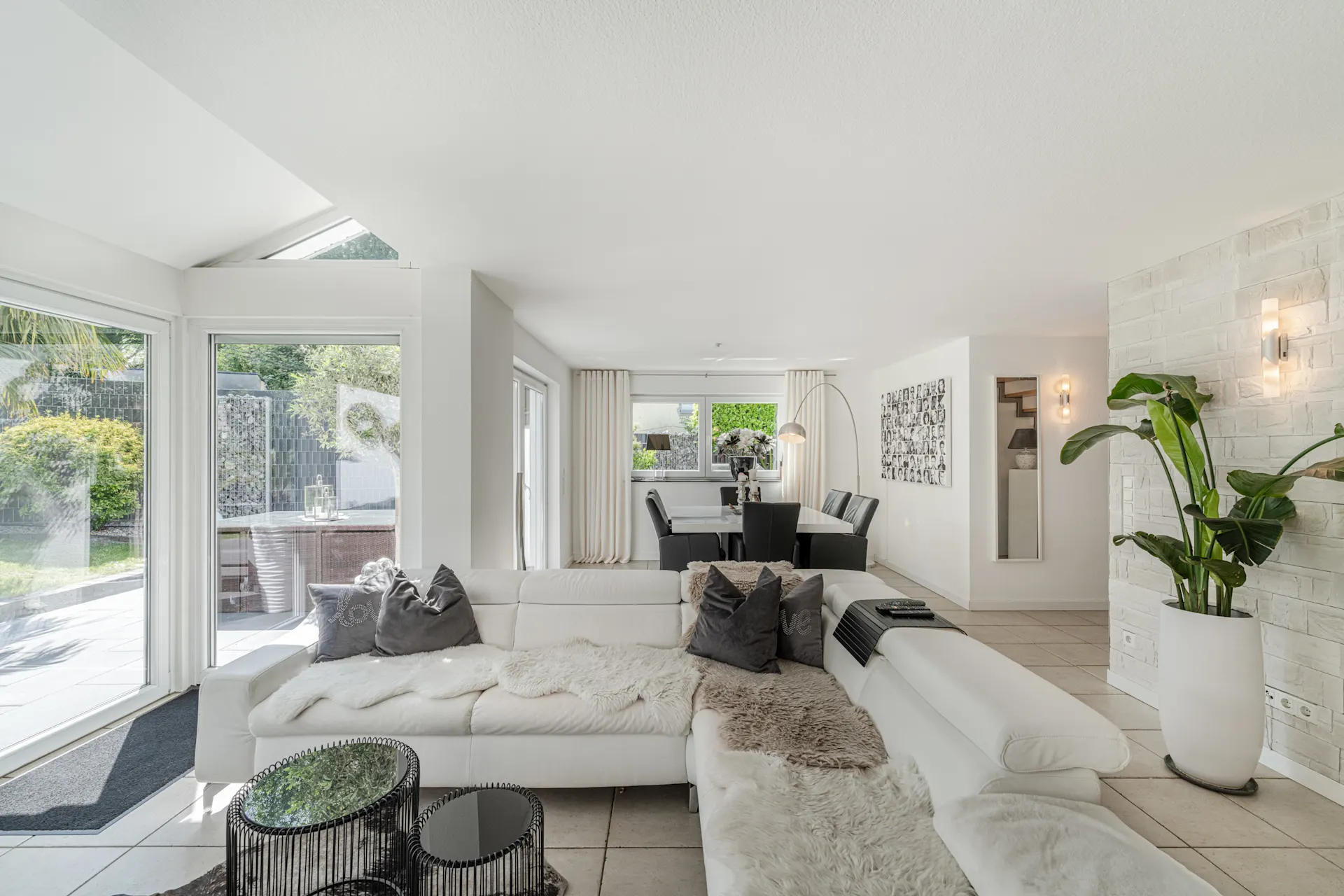 Helles Wohnzimmer mit weißem Sofa, Glastischen, Essbereich und großen Fenstern zum Garten.