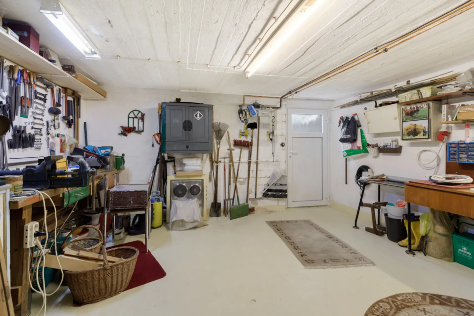 Heller Werkraum mit Werkzeugen an der Wand, Arbeitstischen und Regalen, Türe im Hintergrund.