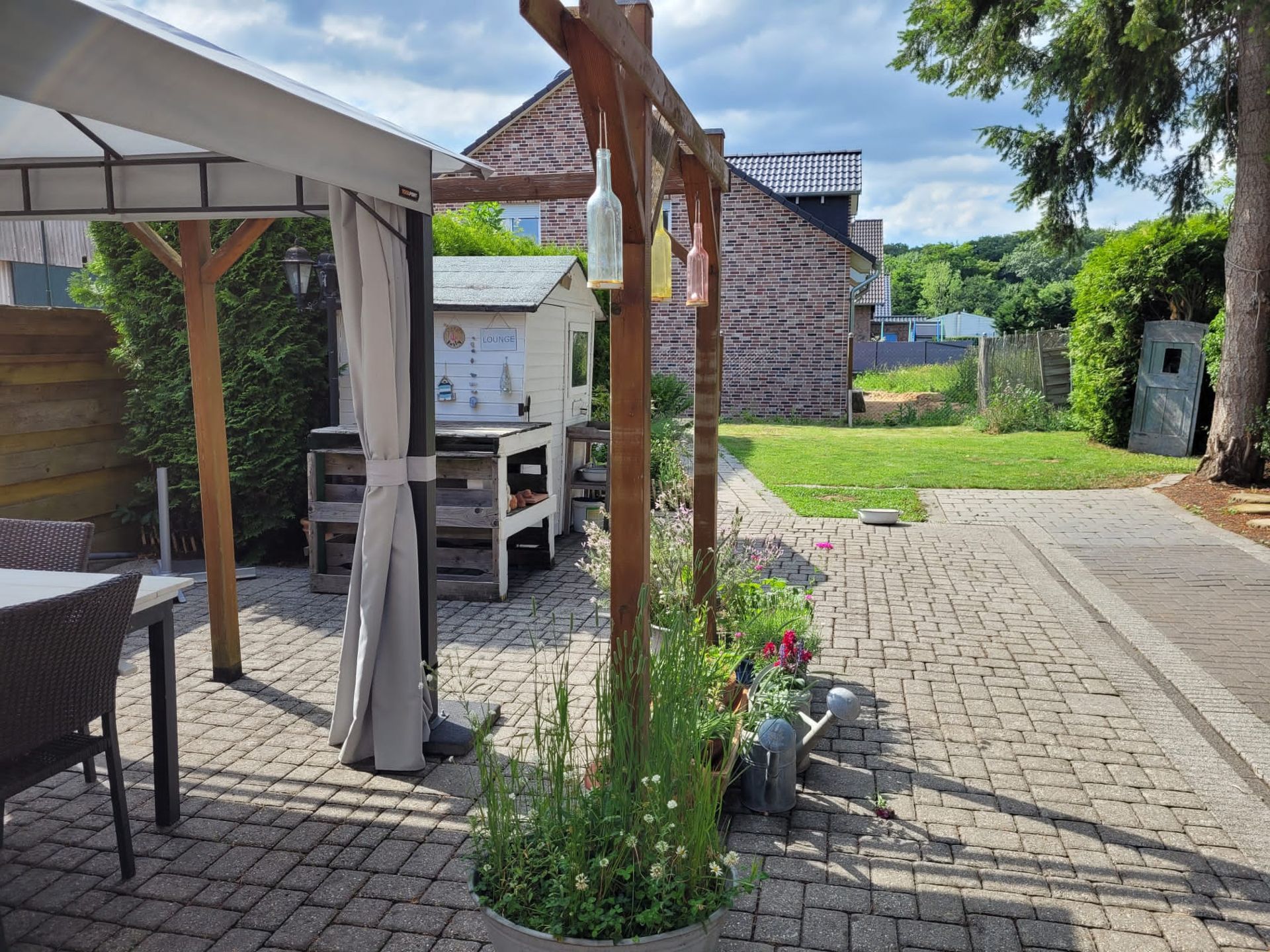 Gepflasterte Terrasse mit Pavillon, Gartenmöbeln und kleinem Gartenhaus im Hintergrund.