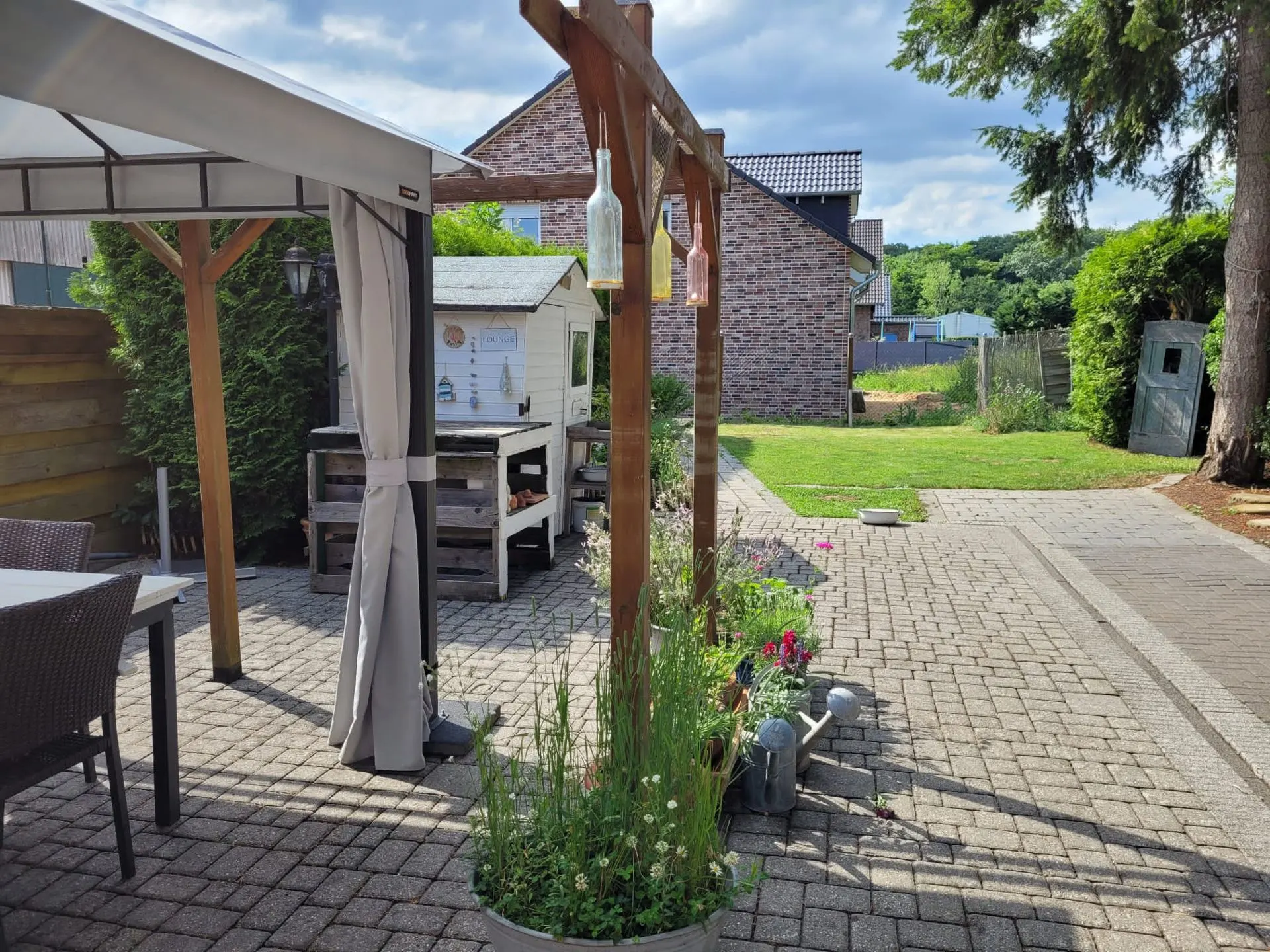 Gepflasterte Terrasse mit Pavillon, Gartenmöbeln und kleinem Gartenhaus im Hintergrund.