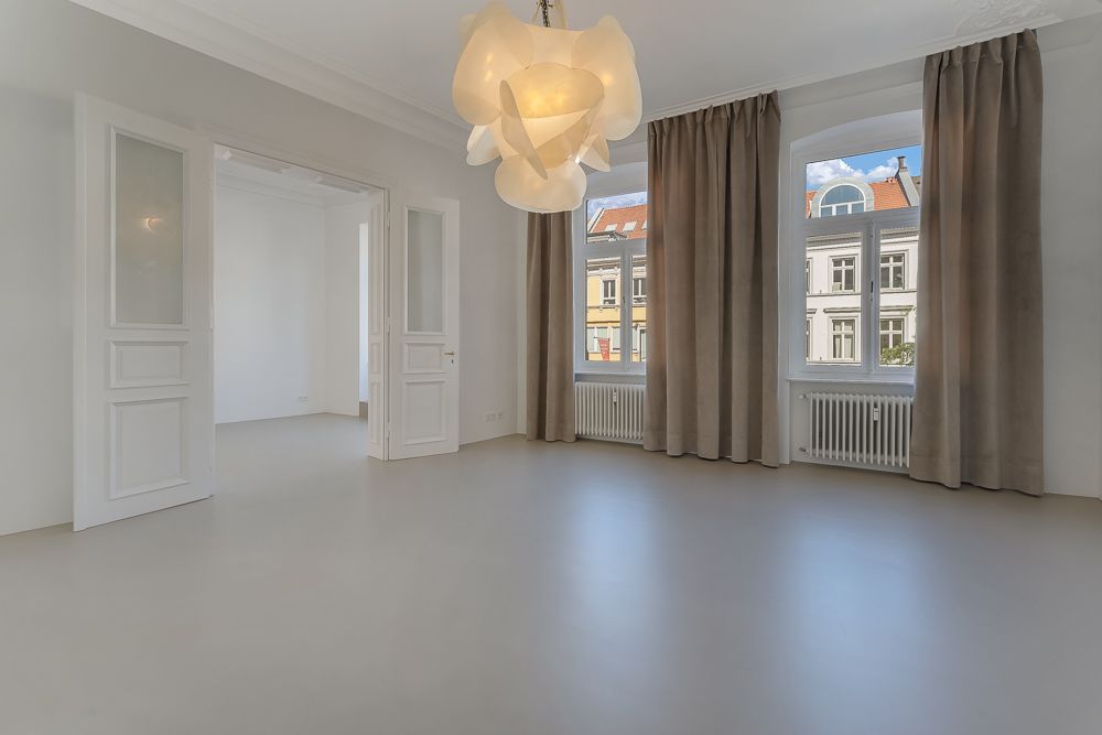 Helles, leeres Zimmer mit drei Fenstern, Vorhängen und einer modernen Deckenlampe.