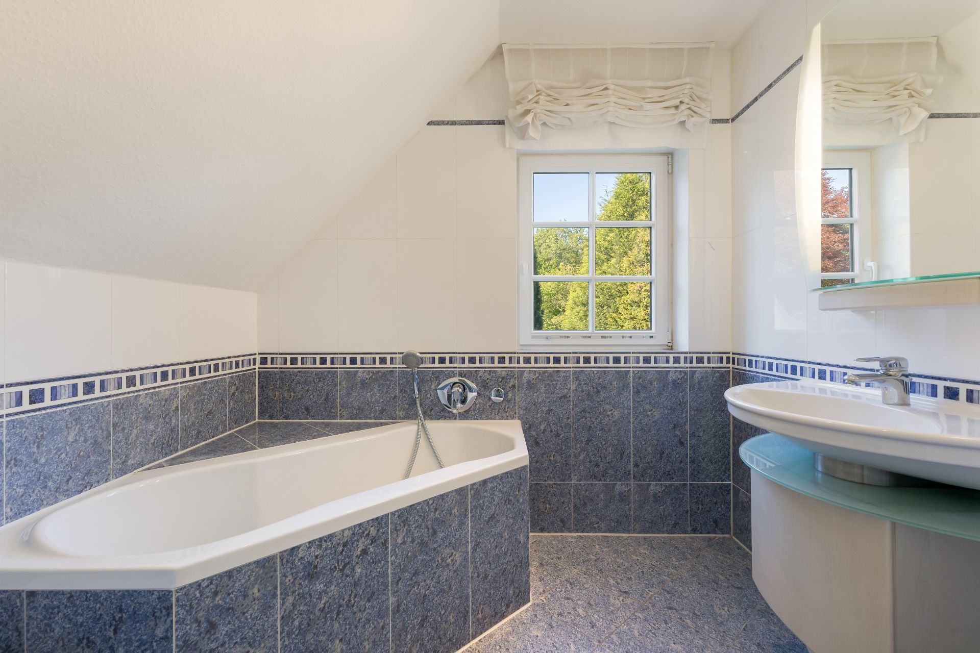 Badezimmer mit Eckbadewanne, Waschbecken und Fenster mit Blick ins Grüne.