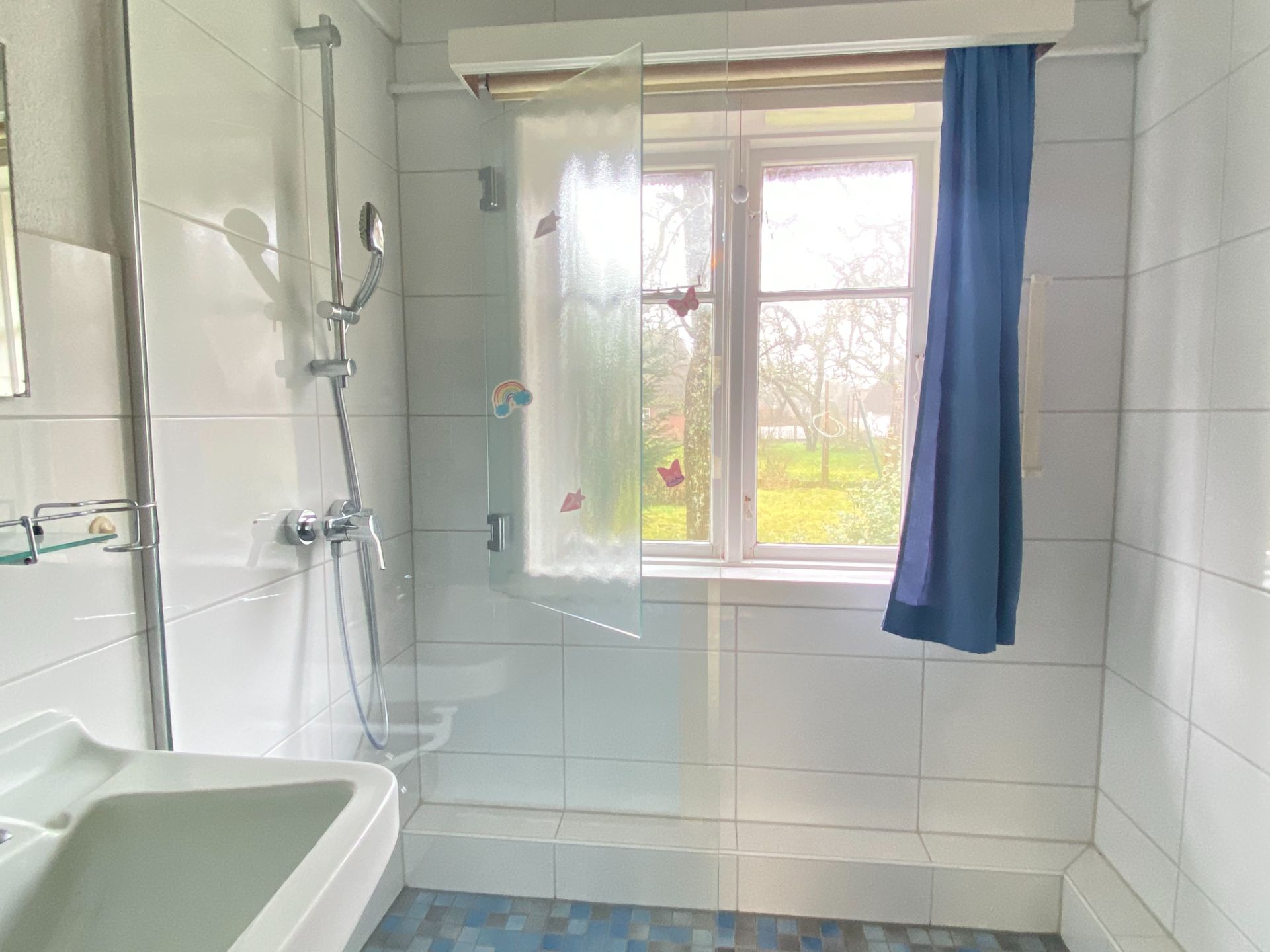 Helles Badezimmer mit Dusche, Waschbecken und Fenster mit blauem Vorhang.