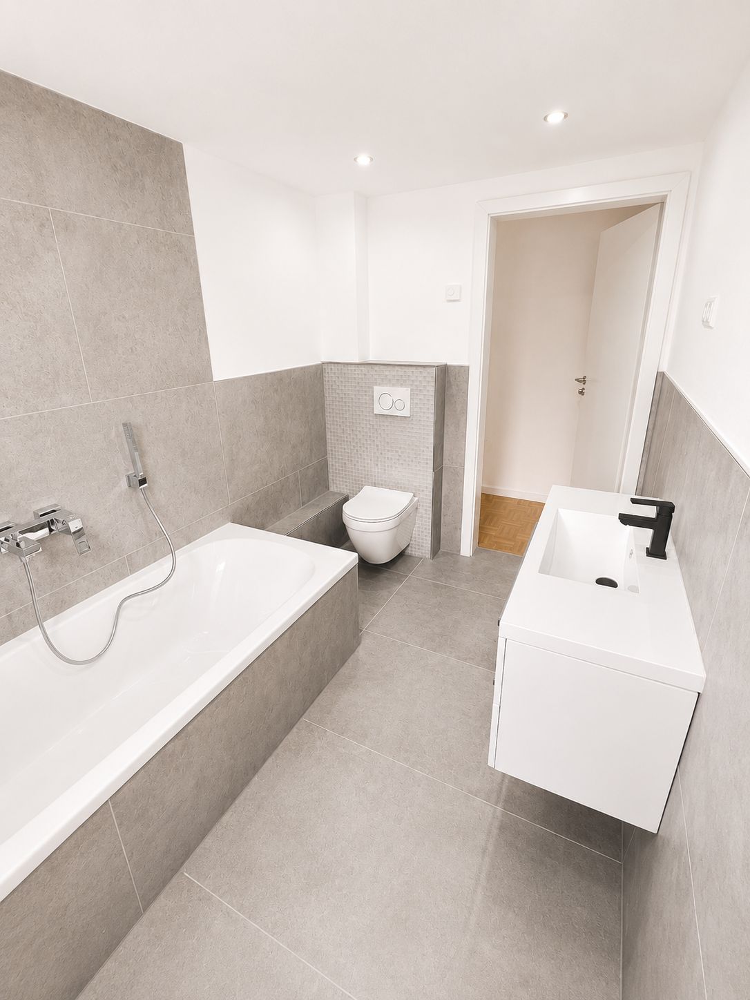 Modernes Badezimmer mit Badewanne, Wandtoilette und Waschbecken auf grauen Fliesen.