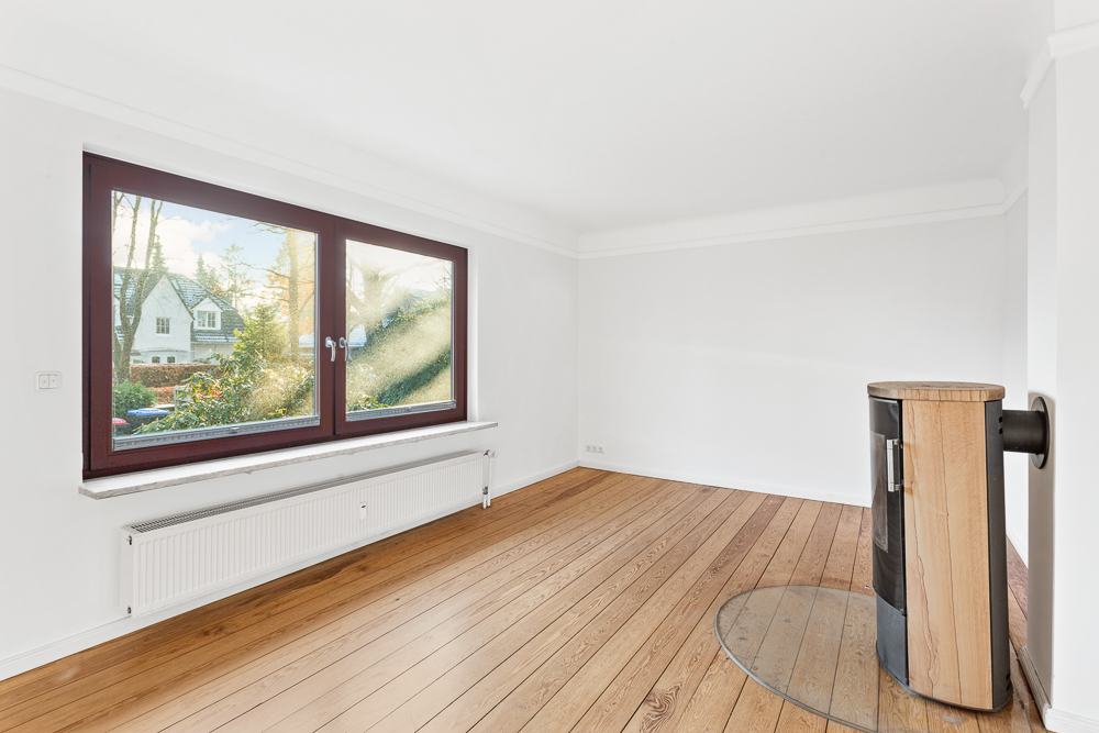 Helles, leeres Zimmer mit Holzfußboden, großem Fenster und Kaminofen.