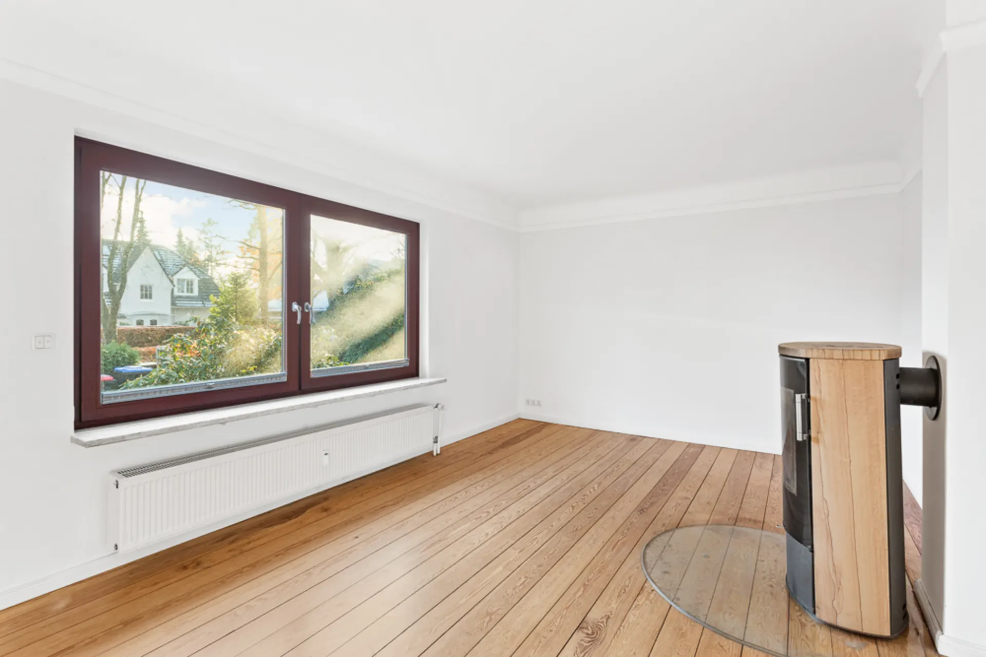 Helles, leeres Zimmer mit Holzfußboden, großem Fenster und Kaminofen.