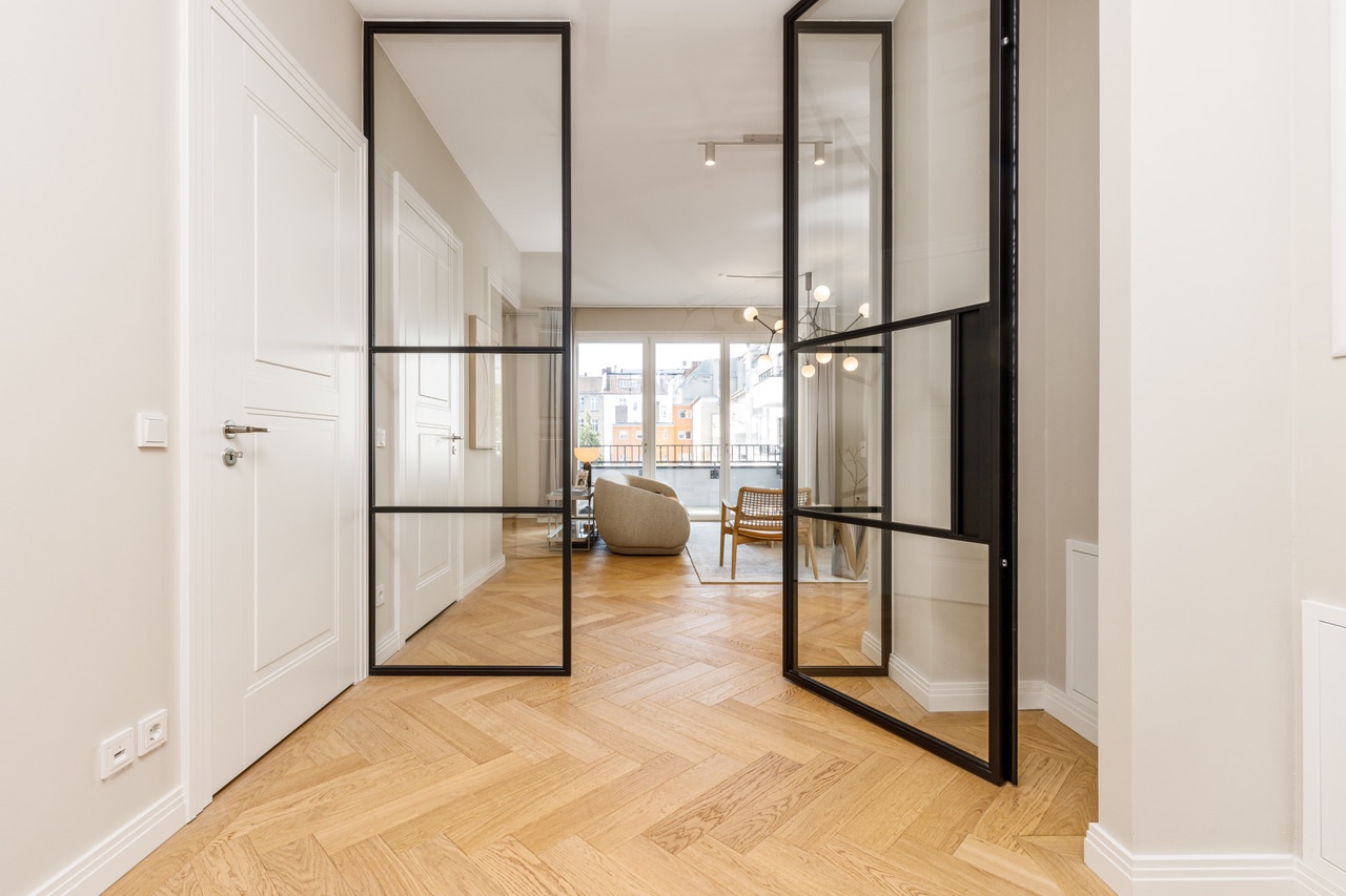 Moderner Flur mit Glastüren, Parkettboden und Blick ins helle Wohnzimmer mit großen Fenstern.