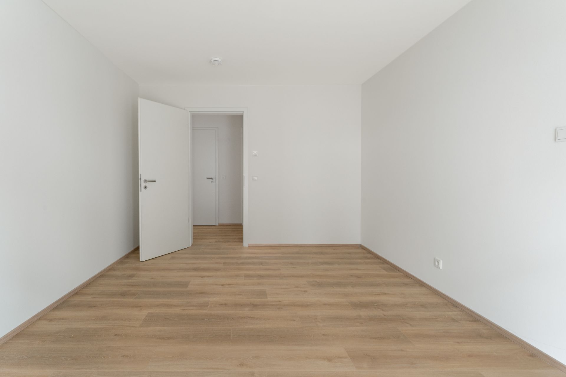 Leerer Raum mit hellem Holzboden und weißen Wänden, offene Tür im Hintergrund.