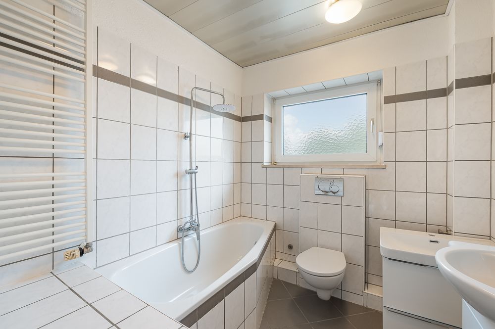 Helles Badezimmer mit Badewanne, Wandheizung, Toilette, Waschbecken und Fenster.