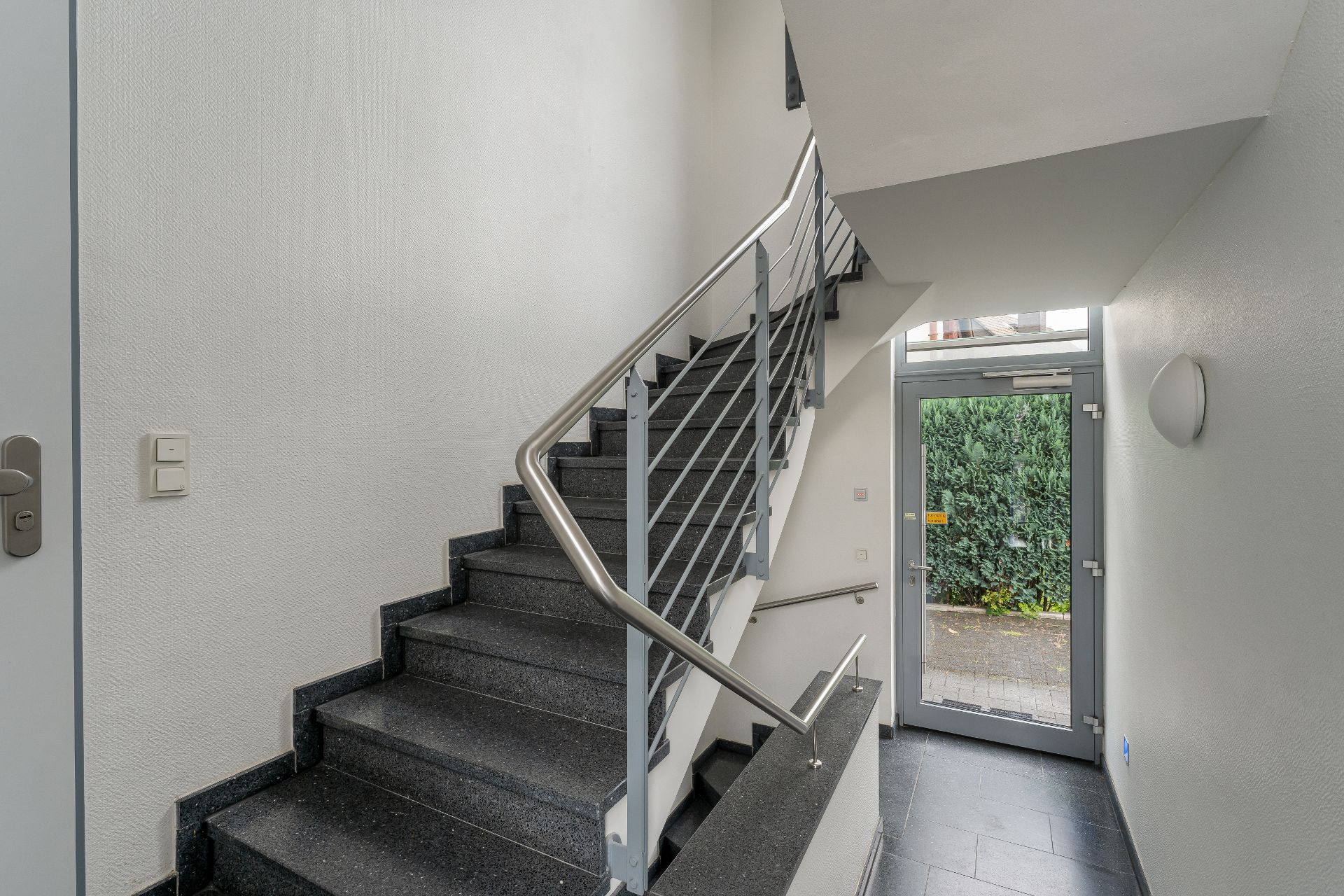 Treppenhaus mit dunklen Stufen und Edelstahlgeländer, neben einer Glastür mit Blick ins Freie.