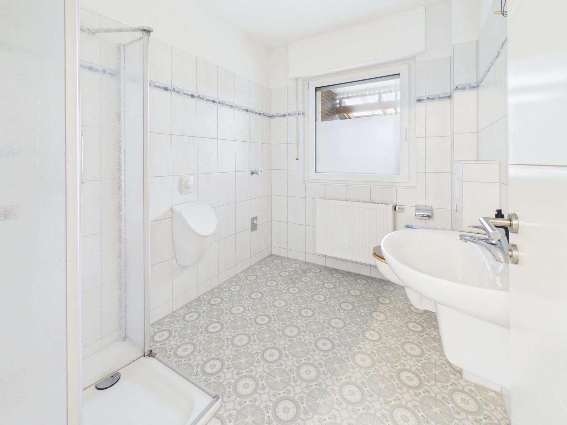 Helles Badezimmer mit Dusche, Waschbecken, Urinal und Fenster.