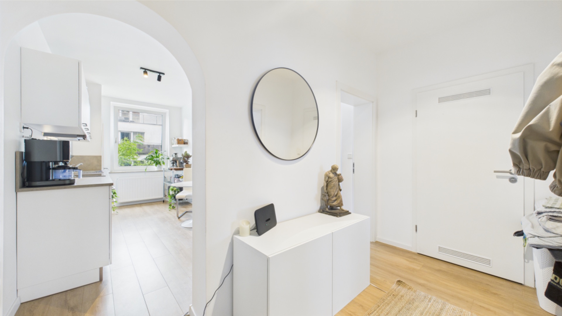 Heller Flur mit rundem Spiegel, weißem Sideboard und Blick in eine Küche mit Fenster.