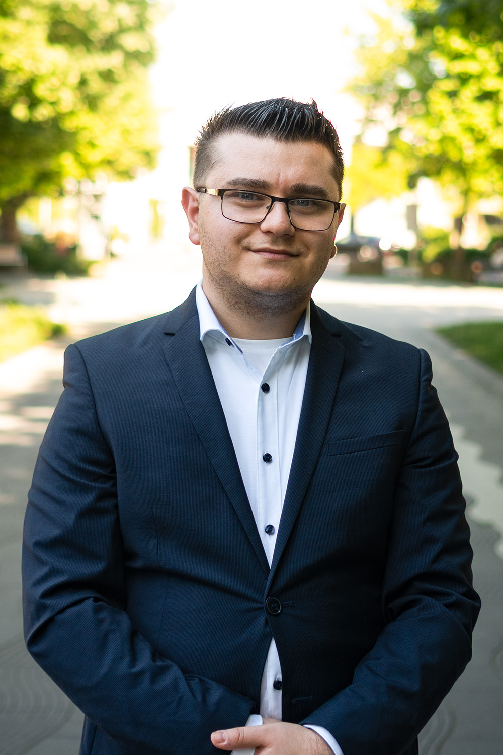 Ein Mann mit Brille im Anzug steht vor Fenstern mit Jalousien.